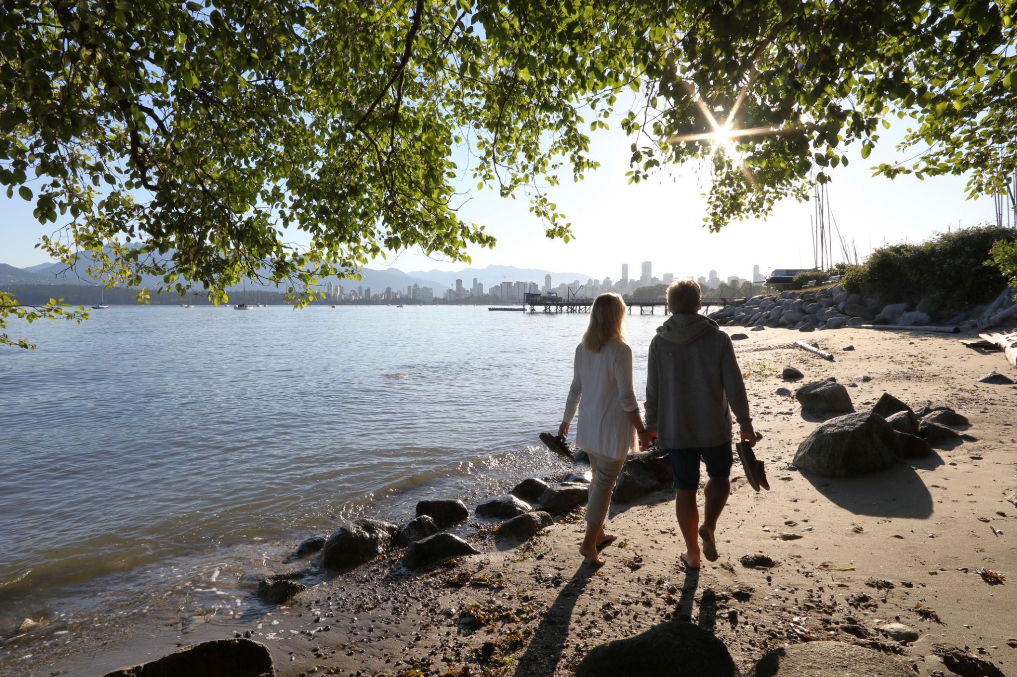 Couple walking along a rocky shoreline in Vancouver with city skyline and mountains in the distance.